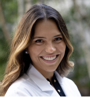A woman with dark brown hair that fades to blonde is wearing a black shirt and a white lab coat while smiling in front of a blurred background of trees.