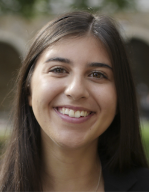 A woman with brown hair is smiling in front of a blurred background.