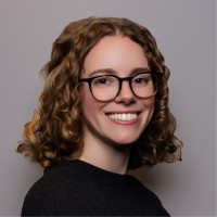 A woman with light, short, curly brown hair is wearing black glasses and a black sweater and smiling. 