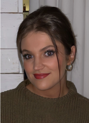 A woman with dark brown hair is wearing a medium brown sweater and is smiling softly in front of a wall.
