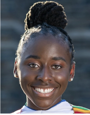 A woman with black hair in a bun and wearing a white, red, yellow, and green shirt is smiling.