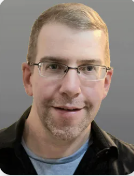A man with short blonde hair and glasses is wearing a a blue shirt and black jacket and is slightly smiling. 