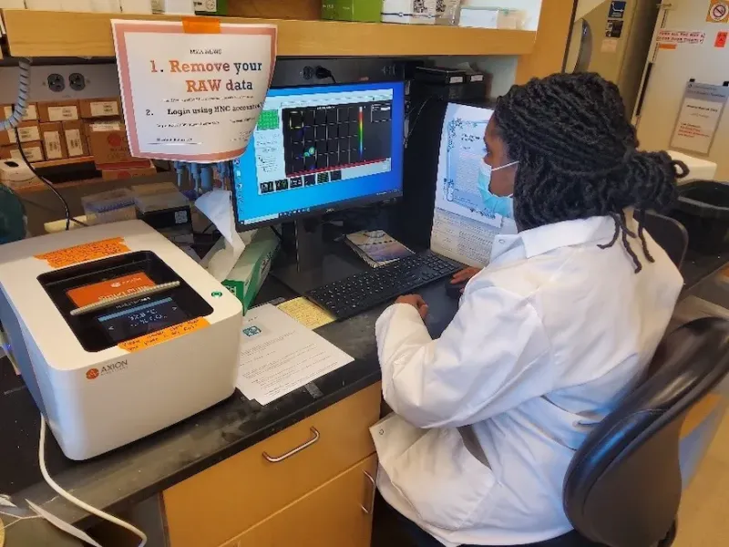 woman in a lab wearing a white lab coat and blue face mask sitting at a desk looking at a computer screen.