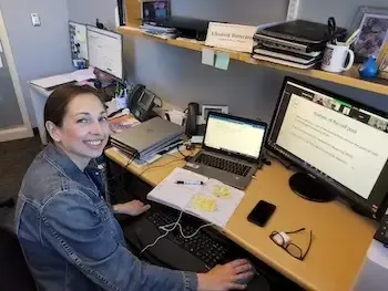 A woman sits at a desk with two computers.