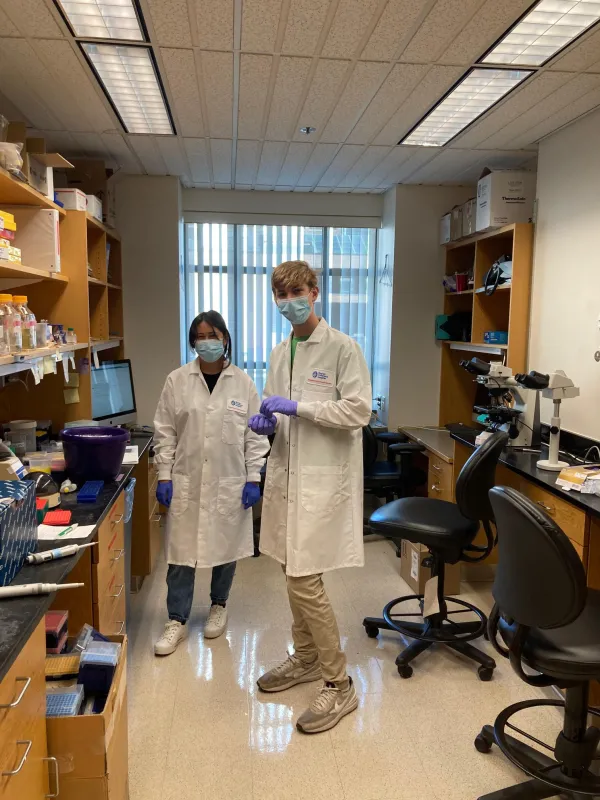 Two interns in the Harris Lab pose in their lab coats, gloves and masks.