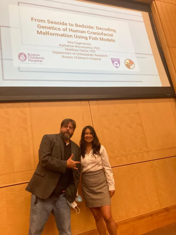 An intern stands with Matthew Harris in front of a presentation sign for decoding genetics of human craniofacial malformation using fish models.