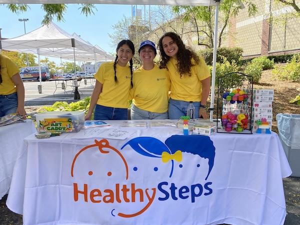 Three women standing behind a table under a tent outside