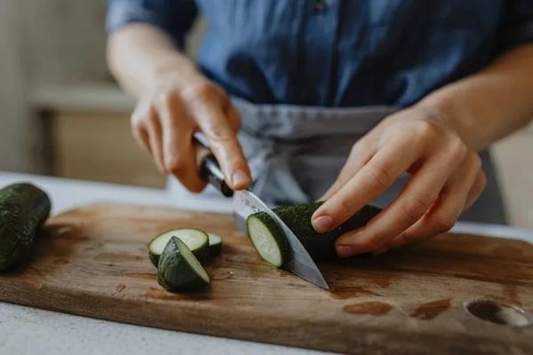 Hands holding a knife while chopping a cucumber on a wooden cutting board