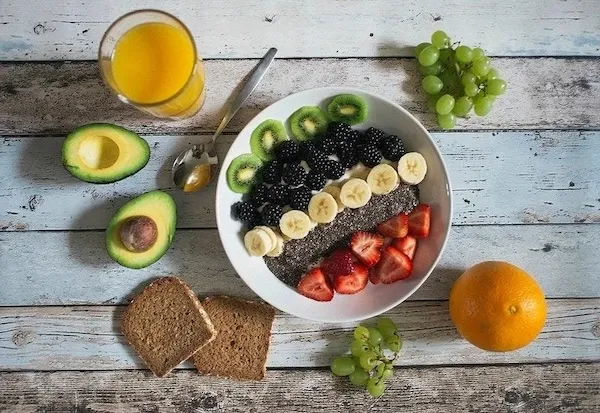 A fruit bowl, orange juice in a glass, wheat toast, grapes, orange, and an avocado sliced in half sitting on a table. 