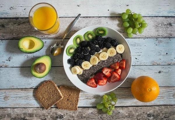 A fruit bowl, orange juice in a glass, wheat toast, grapes, orange, and an avocado sliced in half sitting on a table. 