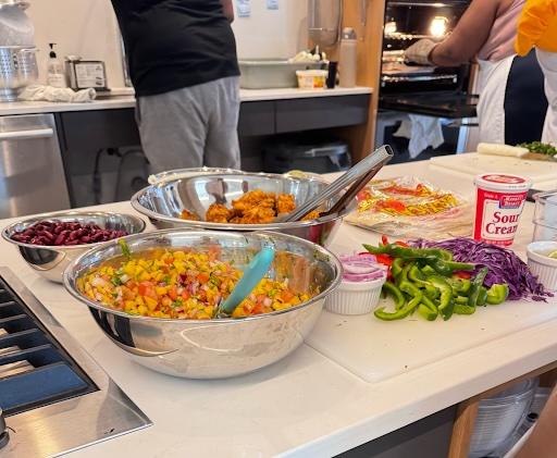 Three bowls of chopped vegetables on a table in a kitchen with two people cooking