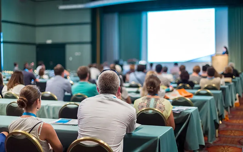 A room full of men and women sitting at tables looking at a powerpoint presentation