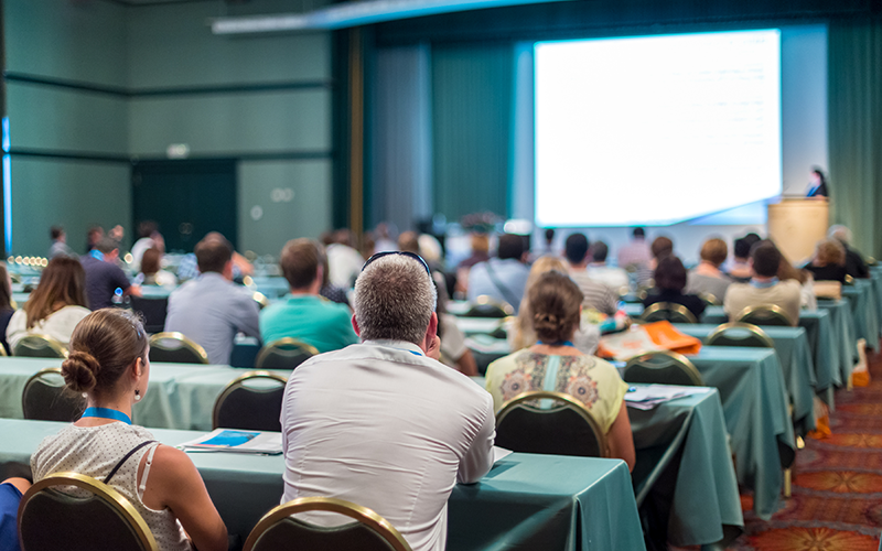 A group of people sit at tables in a large room watching a presentation.