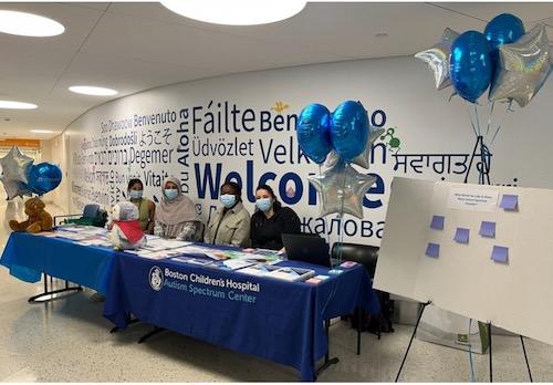 Four women sitting behind tables with blue table cloths with silver and blue balloons and a poster board.