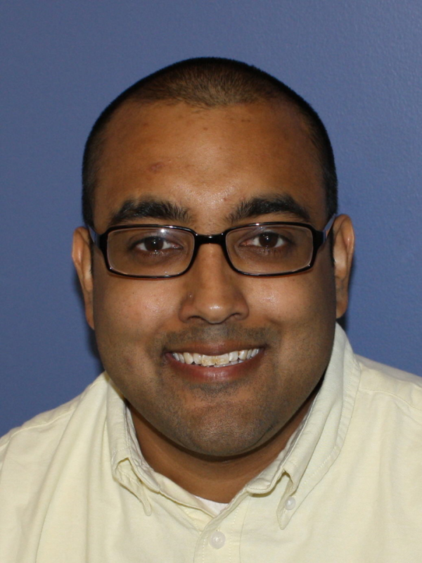 Headshot of Azmar Majmundar, a man with dark skin and a buzzed head wears black square glasses and smiles at the camera.