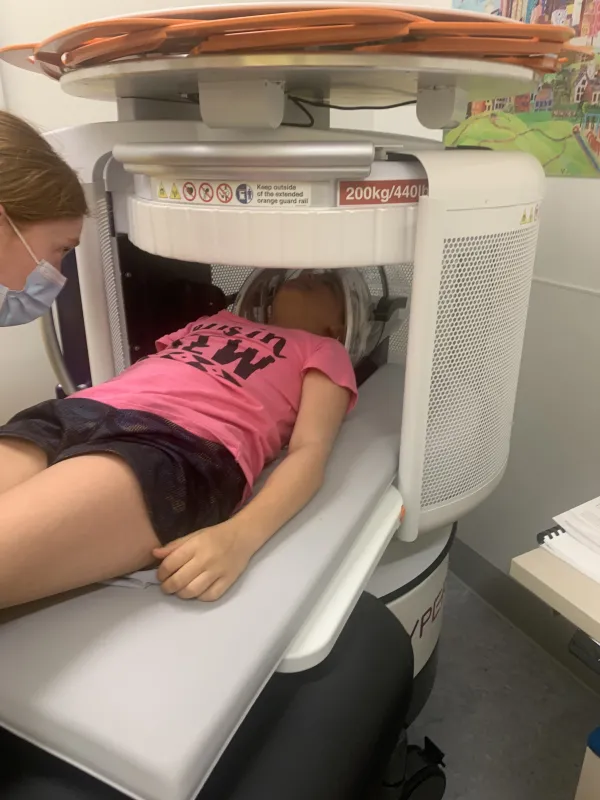 A girl in a pink shirt and black shorts lays in an MRI machine that scans her brain.