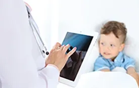 A doctor writes notes on an ipad out of frame for a little boy snuggled up in bed.