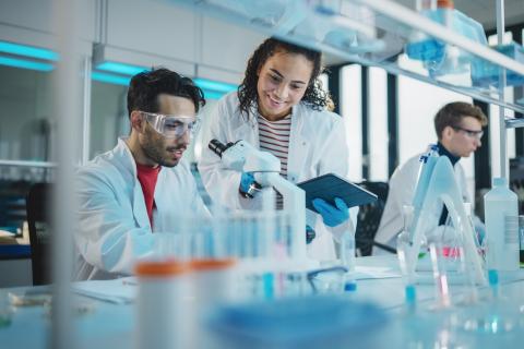 Researcher sits in front of microscope, confers with female colleague