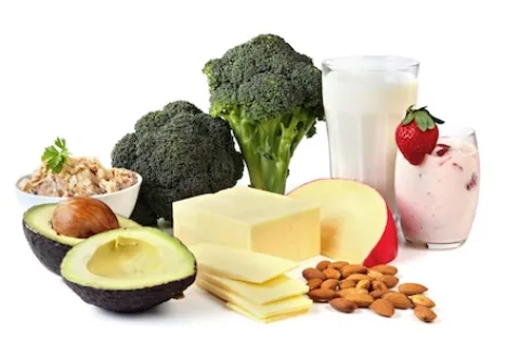 An avocado cut in half, two pieces of broccoli, a block of cheese, almonds, an apple slice, a glass of milk, and a strawberry smoothie.