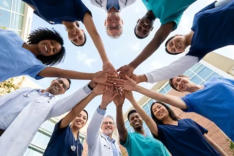 Eleven men and women healthcare providers standing in a circle with hands together in the middle